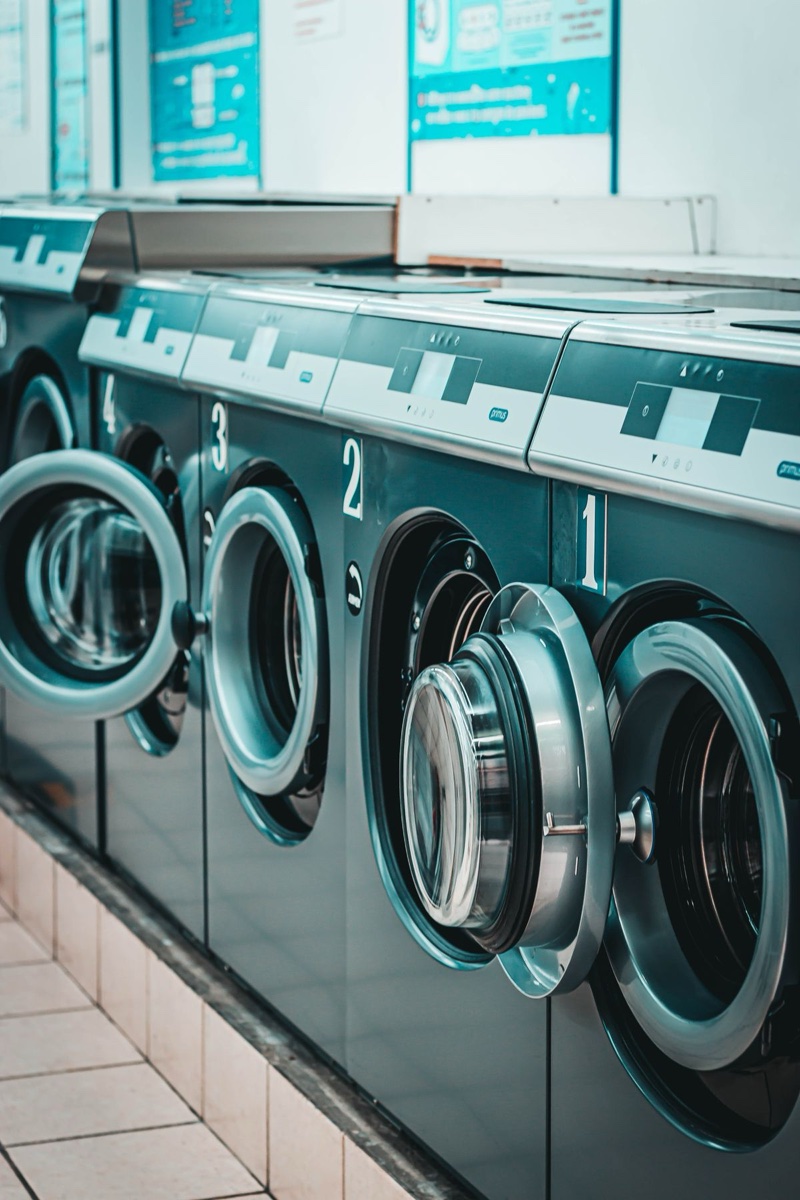Row of numbered industrial washing machines in a laundromat, doors ajar, teal instruction posters on the wall above