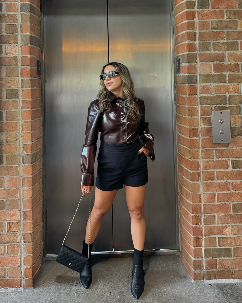 A woman in a glossy burgundy leather jacket and black shorts standing in front of a steel elevator between brick columns
