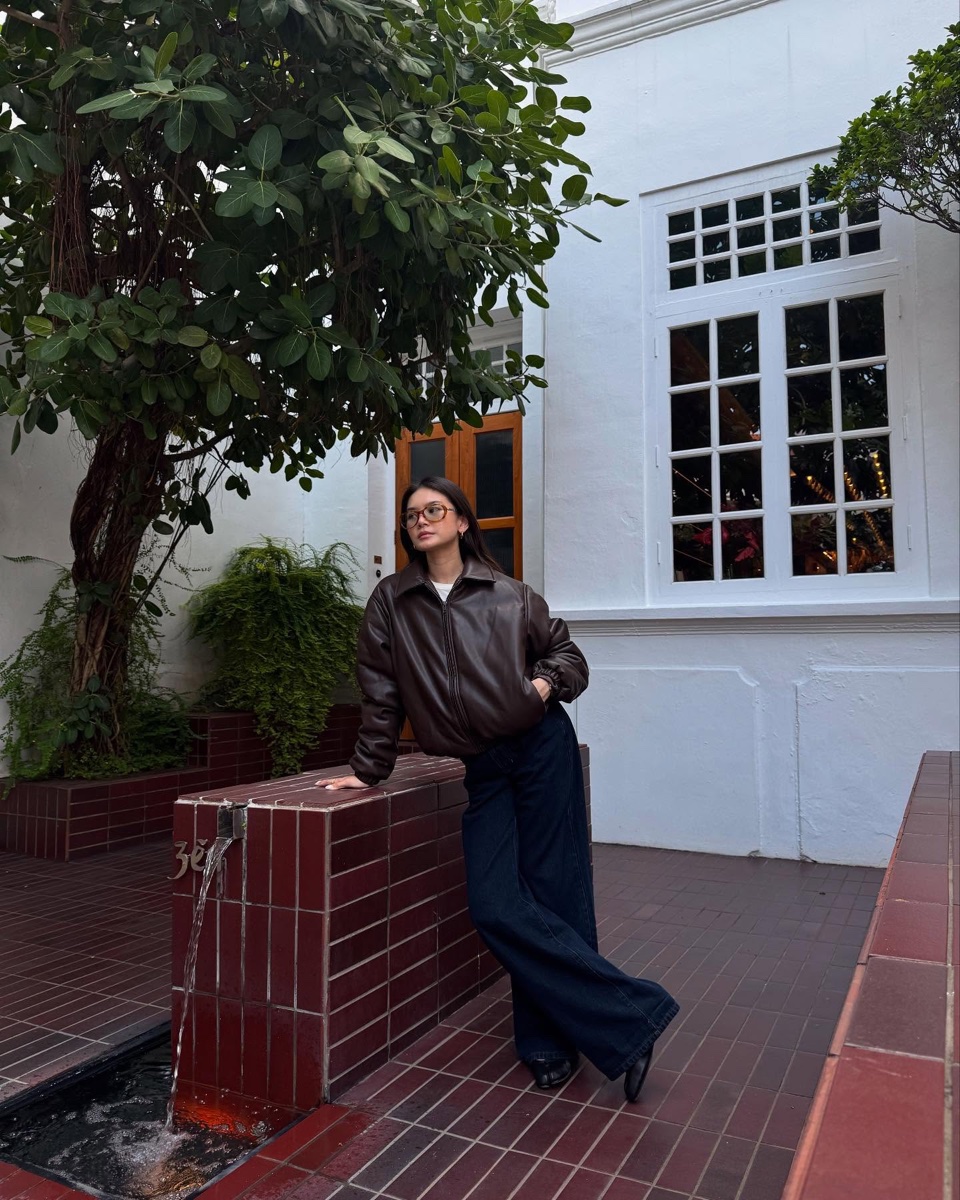 A woman leaning against a tiled courtyard wall in an oversized brown faux leather bomber jacket and wide-leg navy trousers
