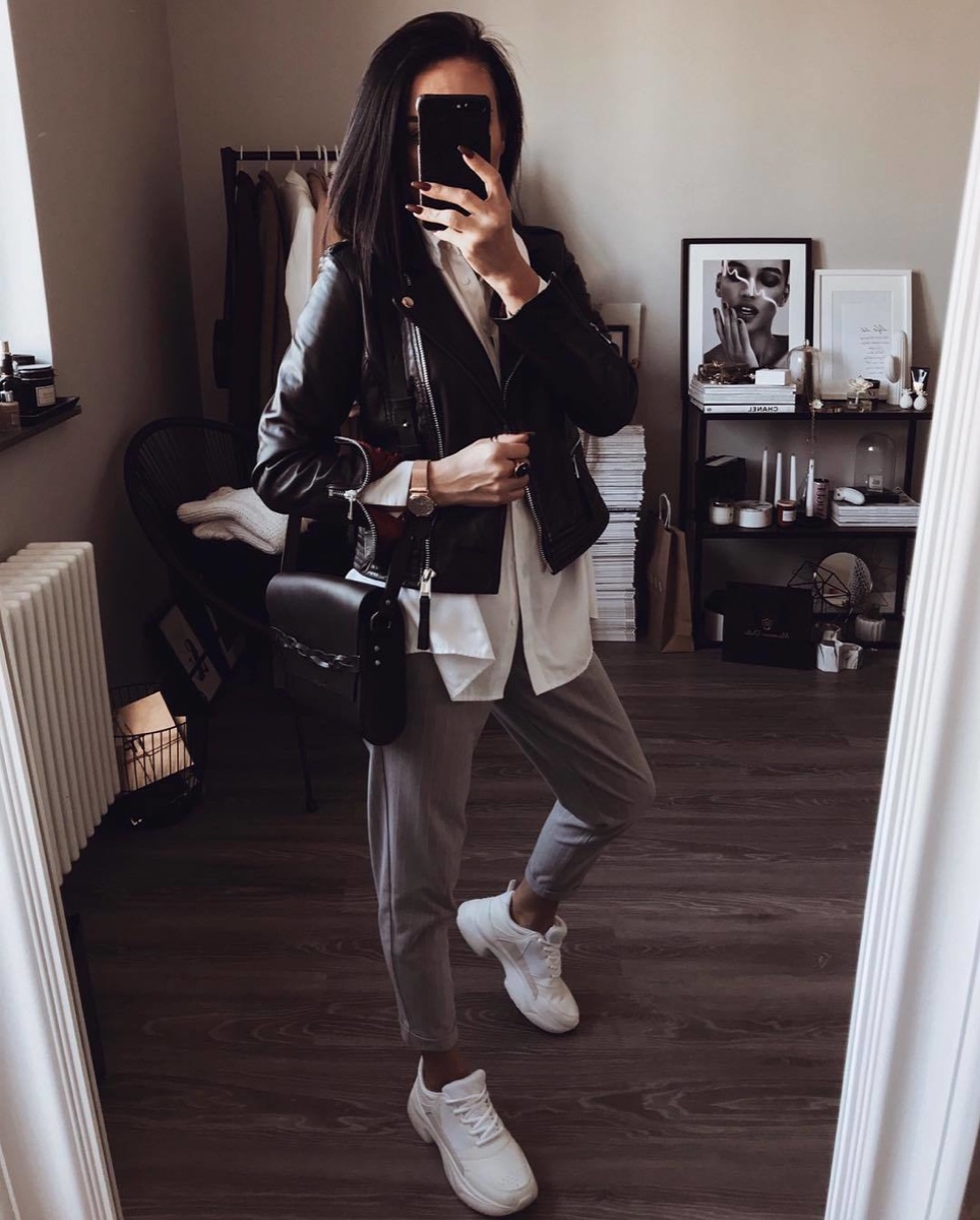 A woman taking a mirror selfie in a black leather jacket over a long white shirt and grey trousers