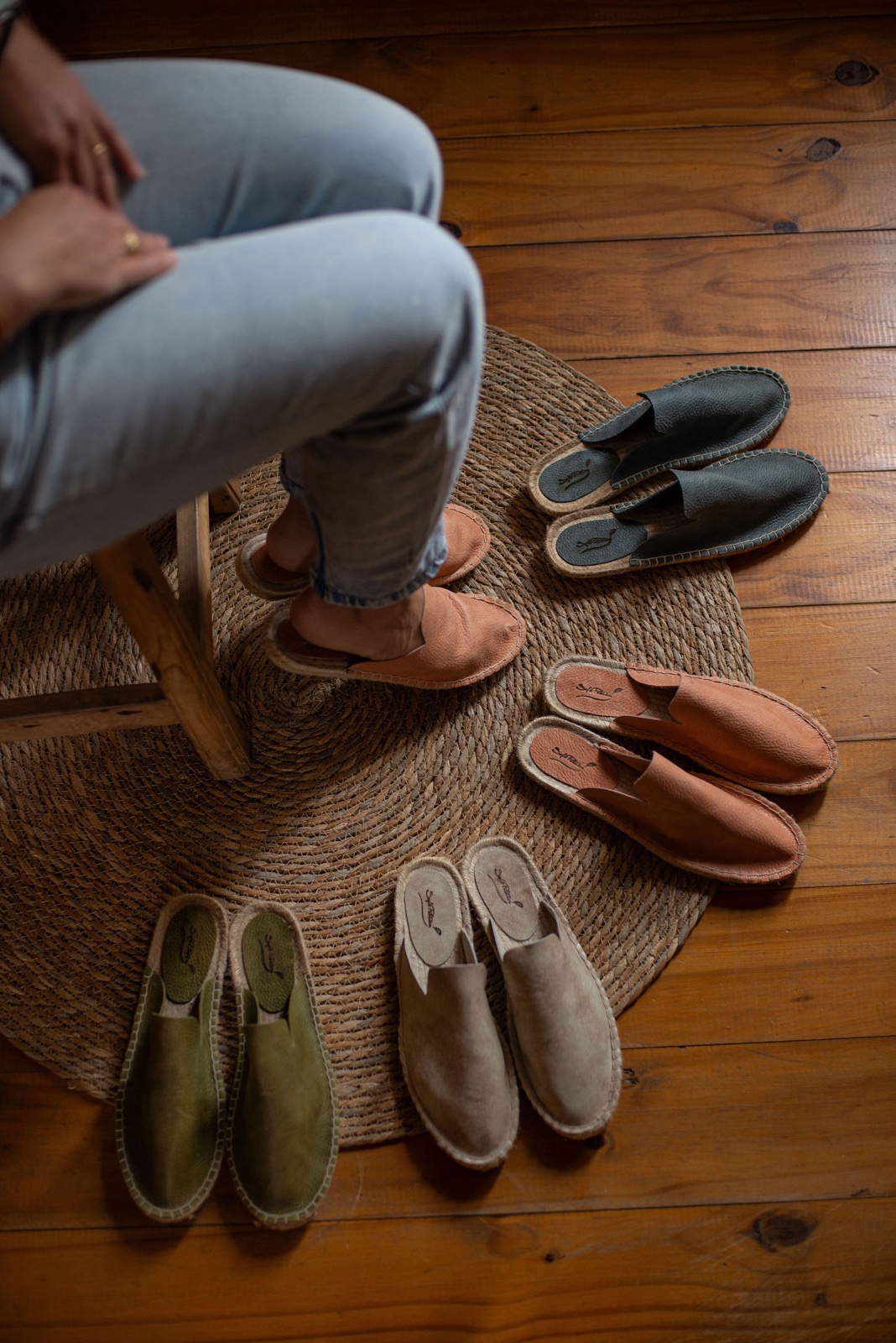 Five pairs of leather slip-on mules in earthy tones arranged on a round woven rug, with a seated person trying on the tan pair
