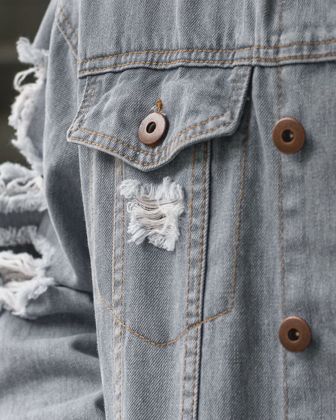 Close-up of a grey denim jacket with copper snaps and frayed openings in the fabric at the sleeve and chest