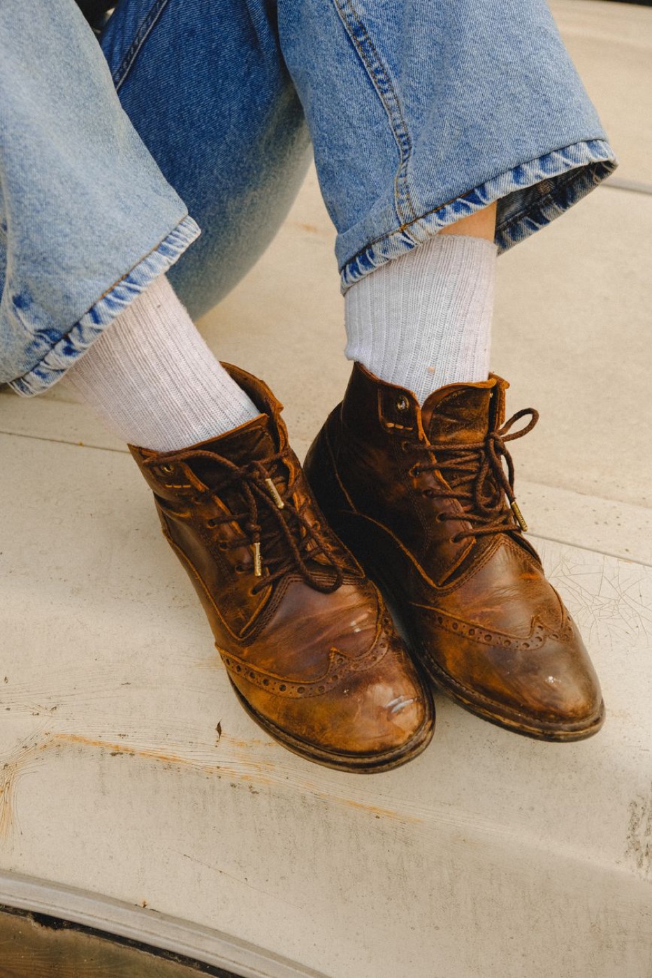 Two feet in white ribbed crew socks and scuffed brown leather brogue boots resting on a concrete step, jeans cuffed above