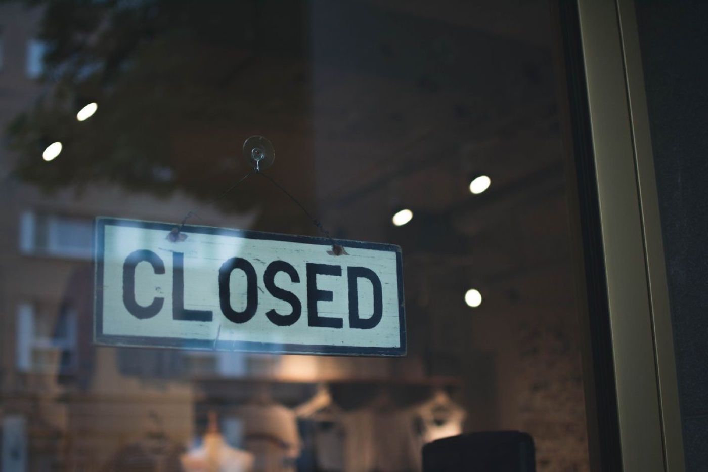 A hand-painted closed sign hanging inside a dark clothing store window with racks visible behind the glass