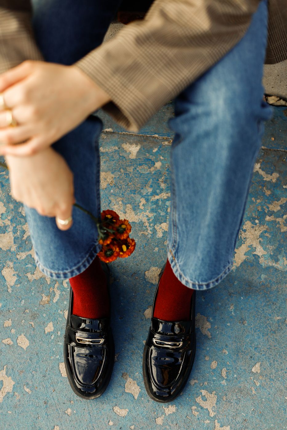 Top-down view of black patent penny loafers worn with dark red socks and straight-leg blue jeans on a weathered blue floor