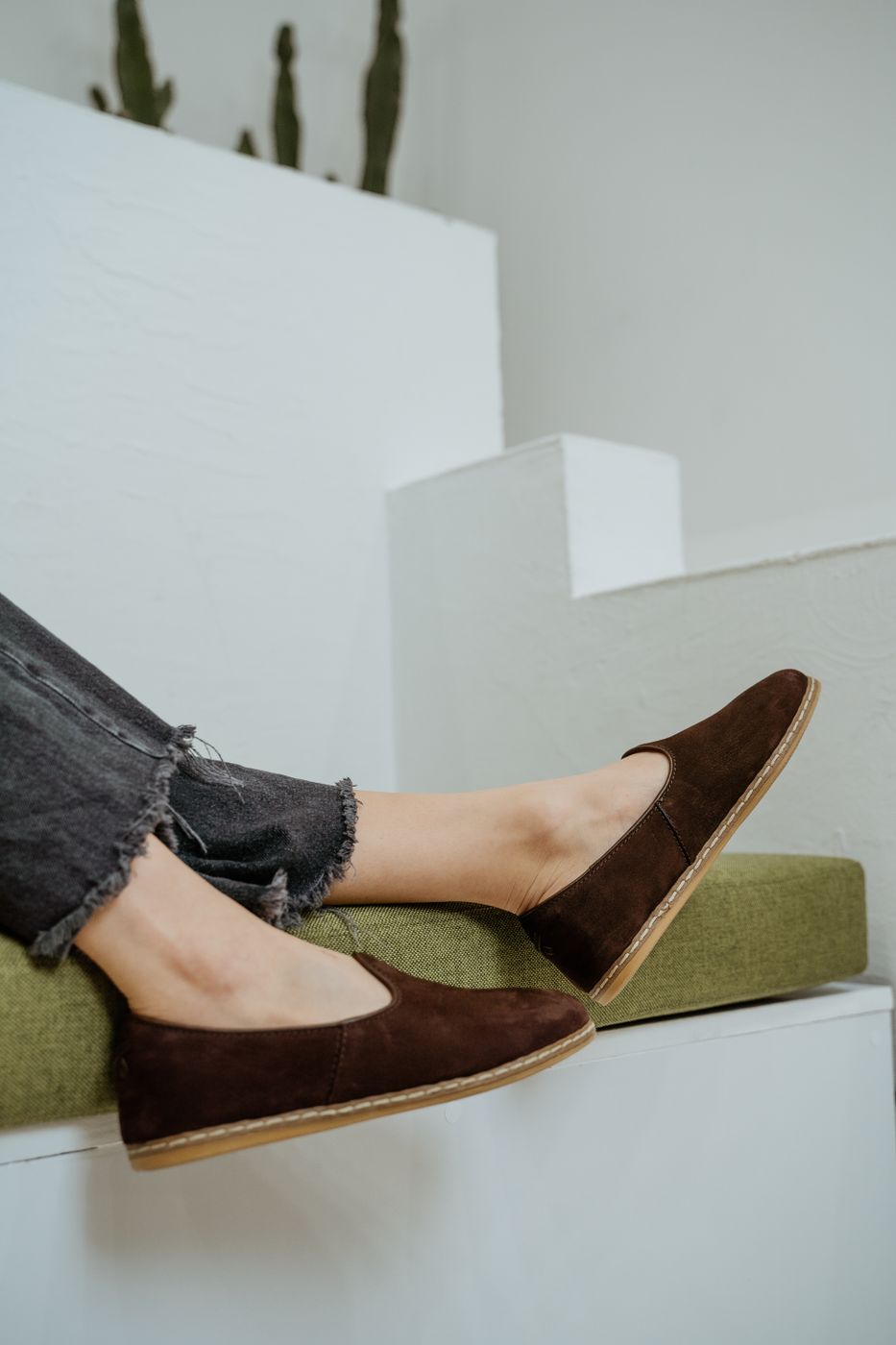 Brown suede soft loafers with an unstructured collapsed profile worn with frayed-hem dark denim against a white stepped wall