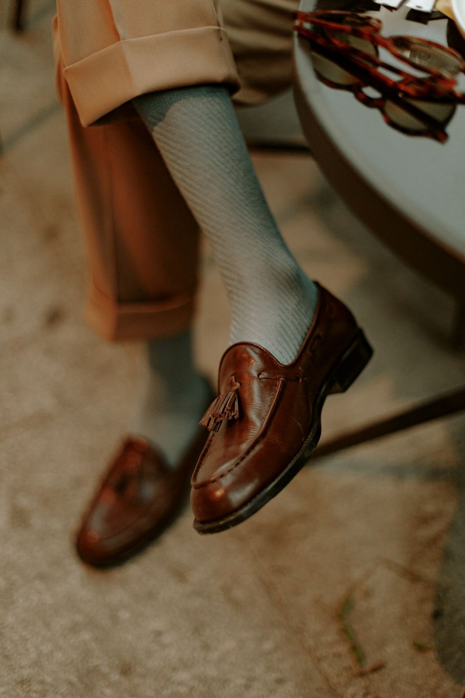 Brown leather tassel loafers worn with pale blue textured knee socks and cuffed camel trousers beside a cafe table