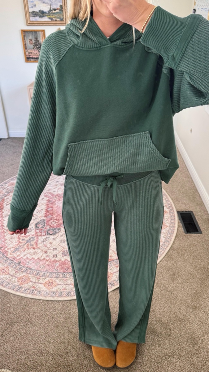 A forest green waffle-knit hoodie and matching drawstring pants worn with tan slippers, standing on a patterned rug at home