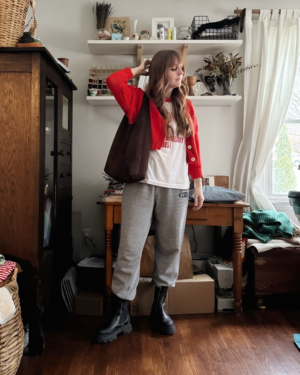 Grey sweatpants worn with a red cardigan, a graphic tee, black lug boots and a brown shoulder bag, in a bedroom with a wooden dresser