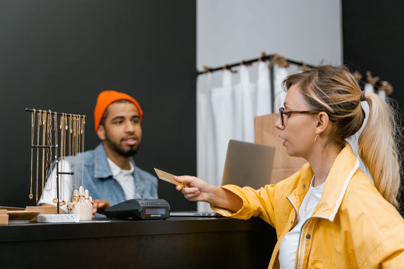 A shopper in a yellow jacket reaches across a dark boutique counter to hand a gold card to a cashier beside a jewelry display