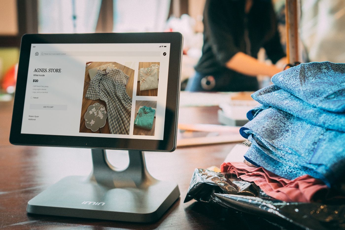 A point-of-sale tablet on a wooden table in a small clothing boutique showing a $120 white hoodie listing, with folded garments stacked beside it