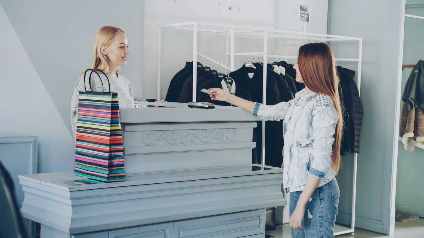 A customer in a denim jacket hands a card across a pale grey checkout counter to a cashier in a clothing store