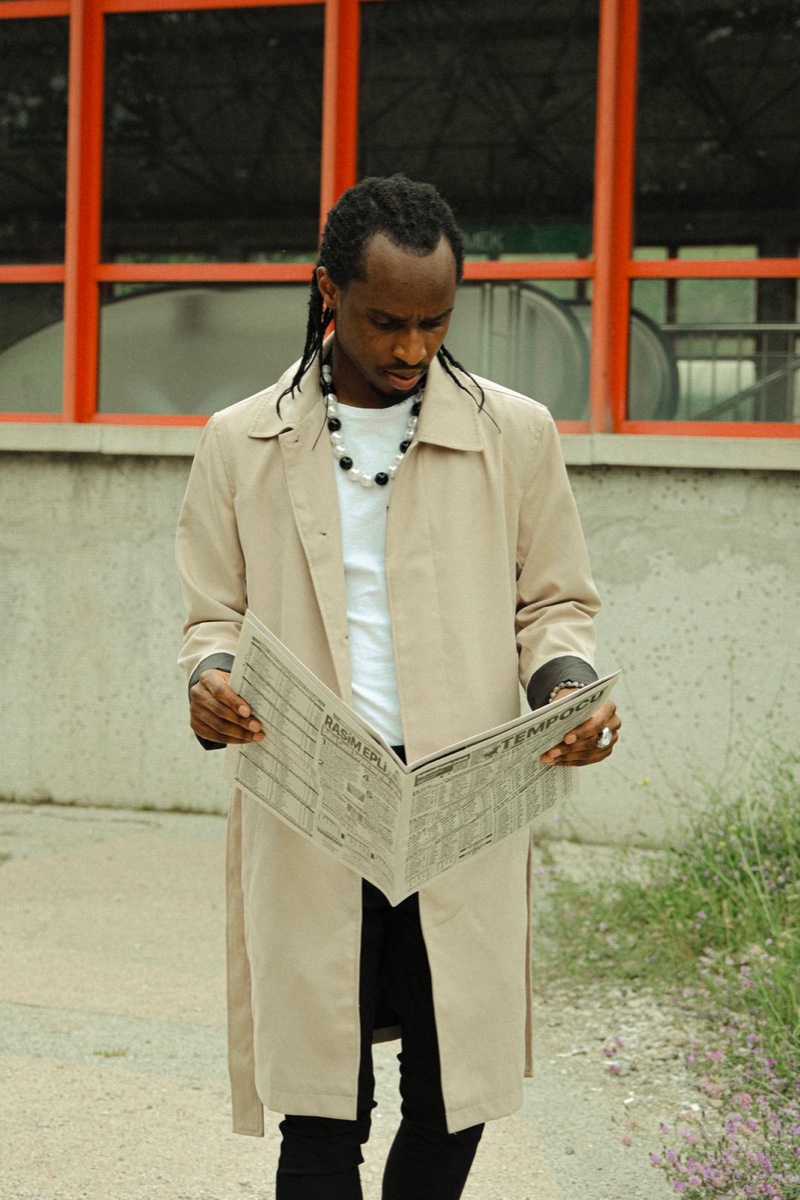 A man in a knee-length beige single-breasted mac coat standing on a cracked pavement reading a newspaper, red-framed industrial windows behind him