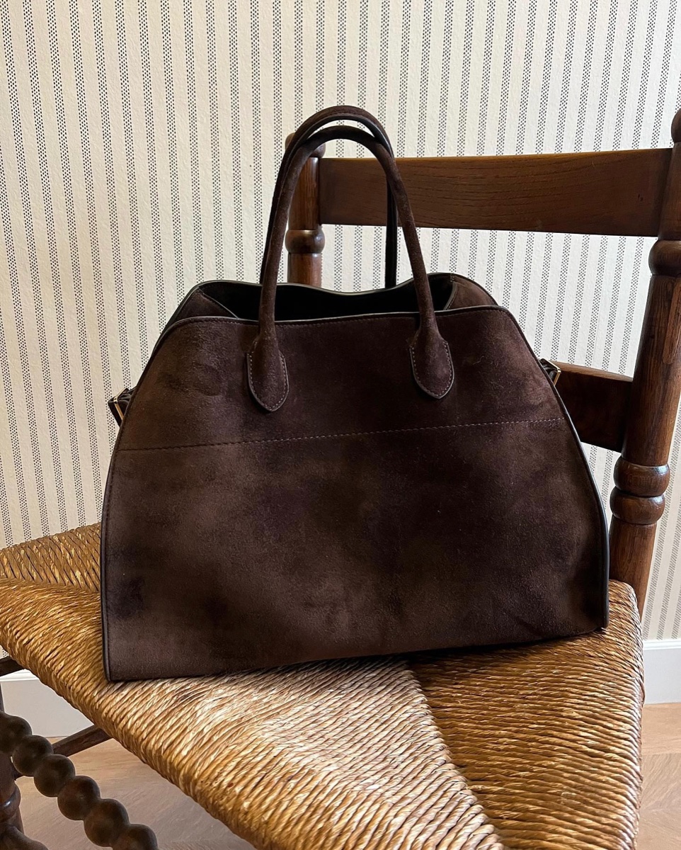 A chocolate brown suede Margaux 15 tote sitting on a rush-seated wooden chair against striped wallpaper