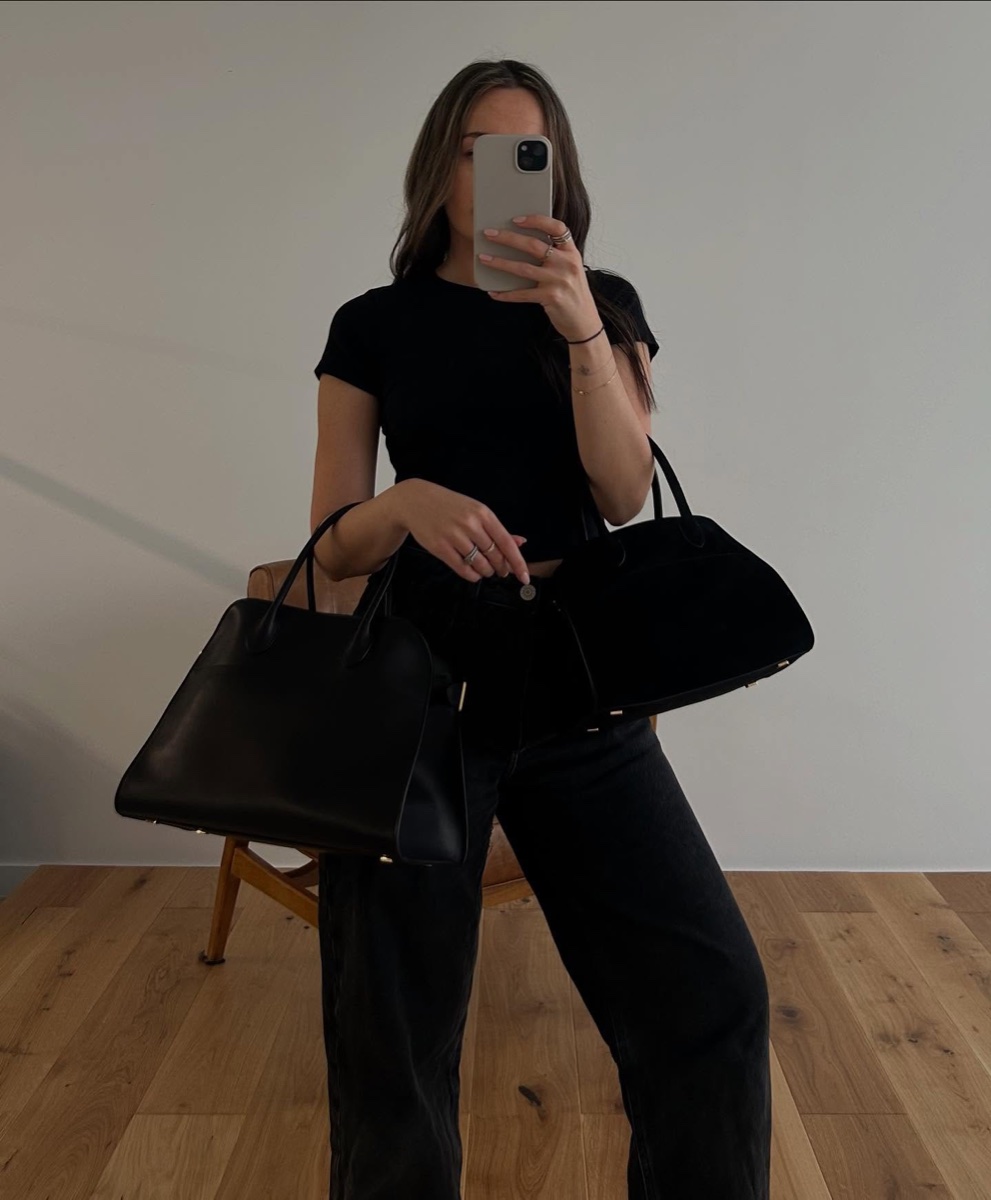A woman in a black t-shirt and black jeans takes a mirror photo holding two Margaux bags, one black leather and one black suede, her face behind the phone