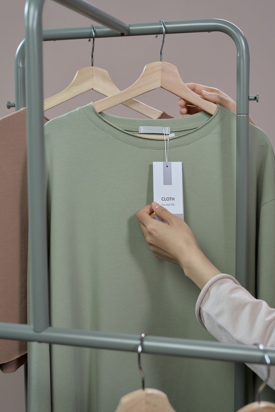 A hand lifting the hang tag on a sage green t-shirt hanging on a metal rail to read what is printed on it