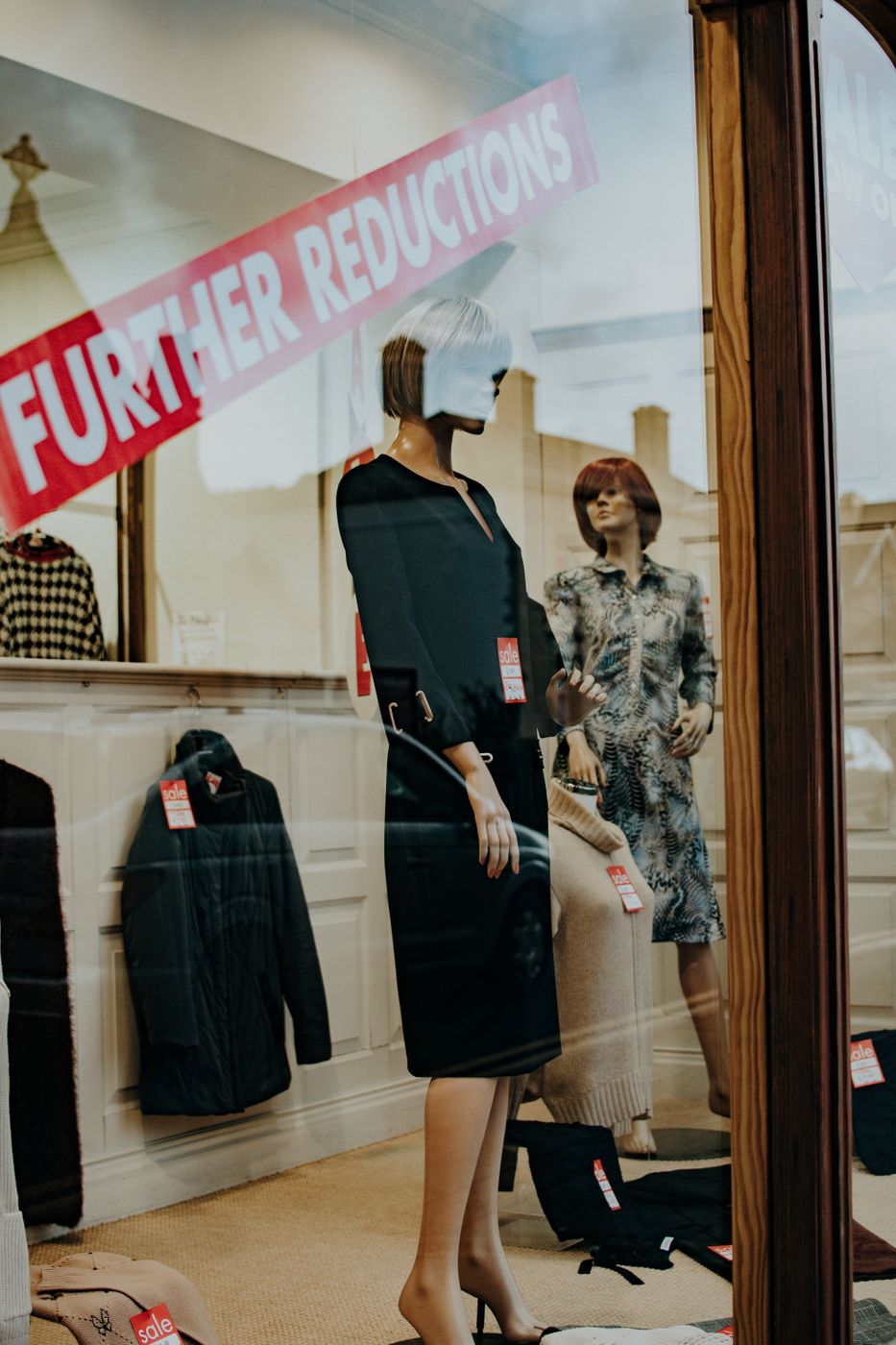 A shop window with a diagonal red FURTHER REDUCTIONS banner across the glass and red sale tags hanging from every garment inside