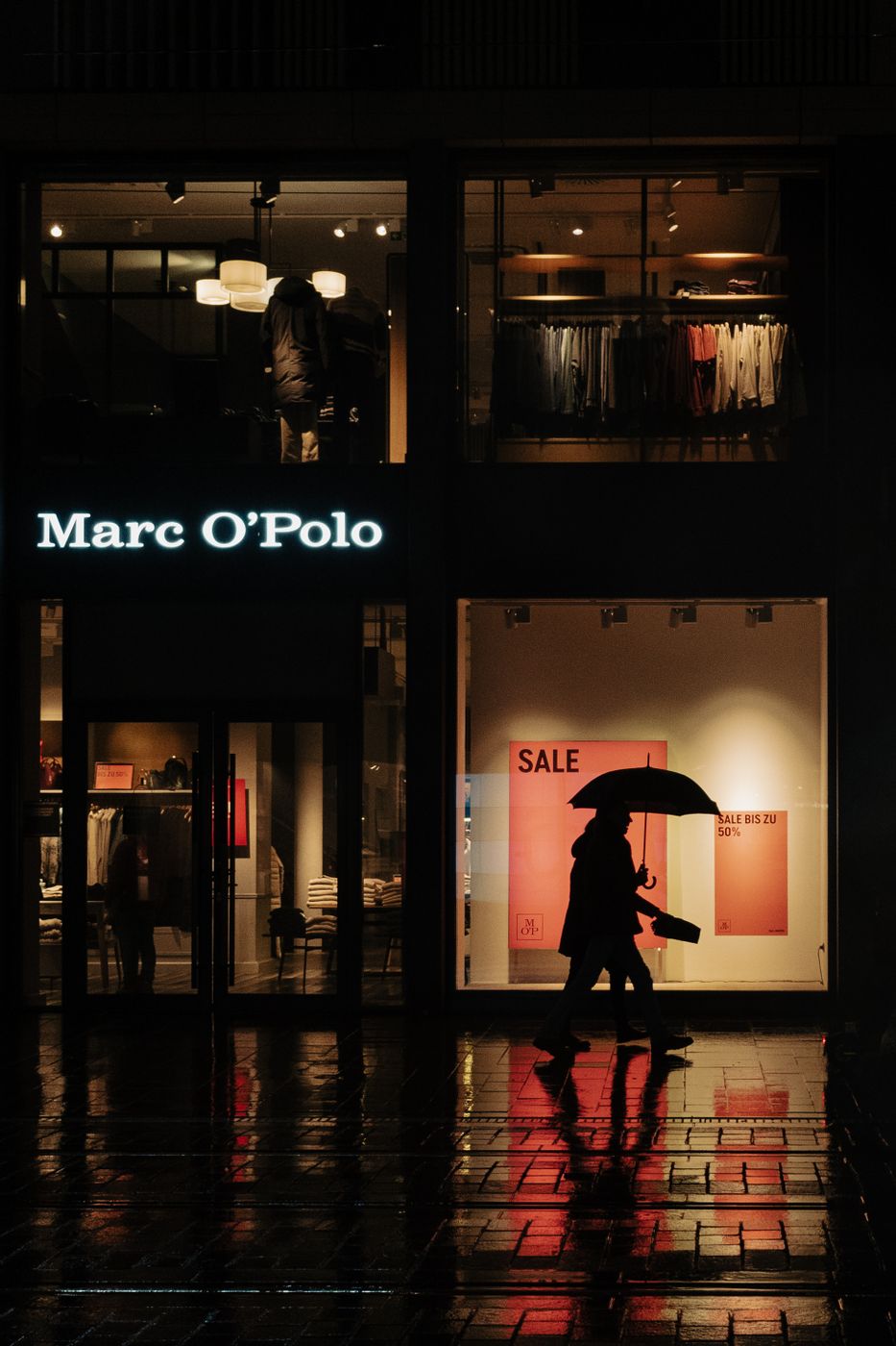 A person with an umbrella walks past a lit clothing store window at night, a red SALE poster glowing behind the glass on a wet pavement