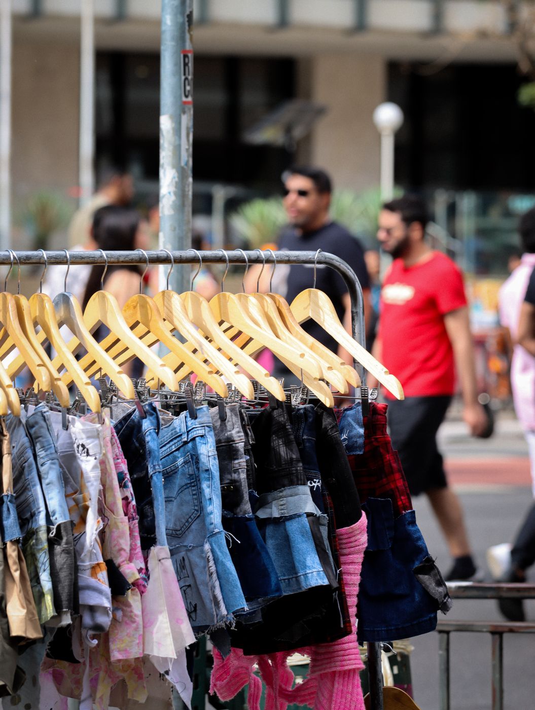 An outdoor market rail of denim shorts and skirts on pale wooden hangers, shoppers walking past out of focus behind it