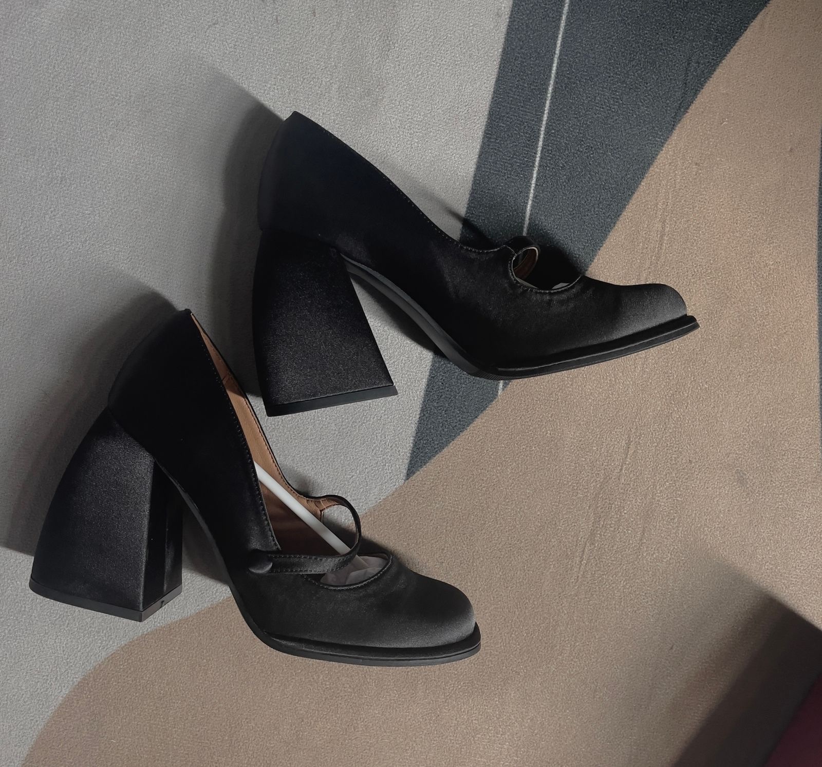 A pair of black satin mary jane pumps with thin straps and flared block heels resting on a patterned rug