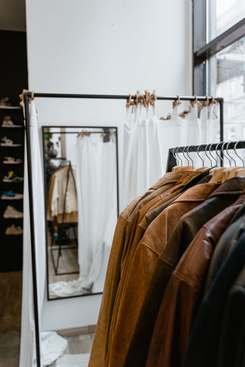 Brown leather and suede jackets hanging on a black steel rack beside a mirror in a shop with window light
