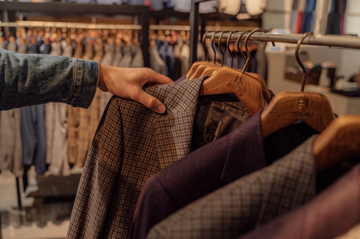 A hand in a denim sleeve pulling a brown checked jacket from a store rack of menswear on wooden hangers