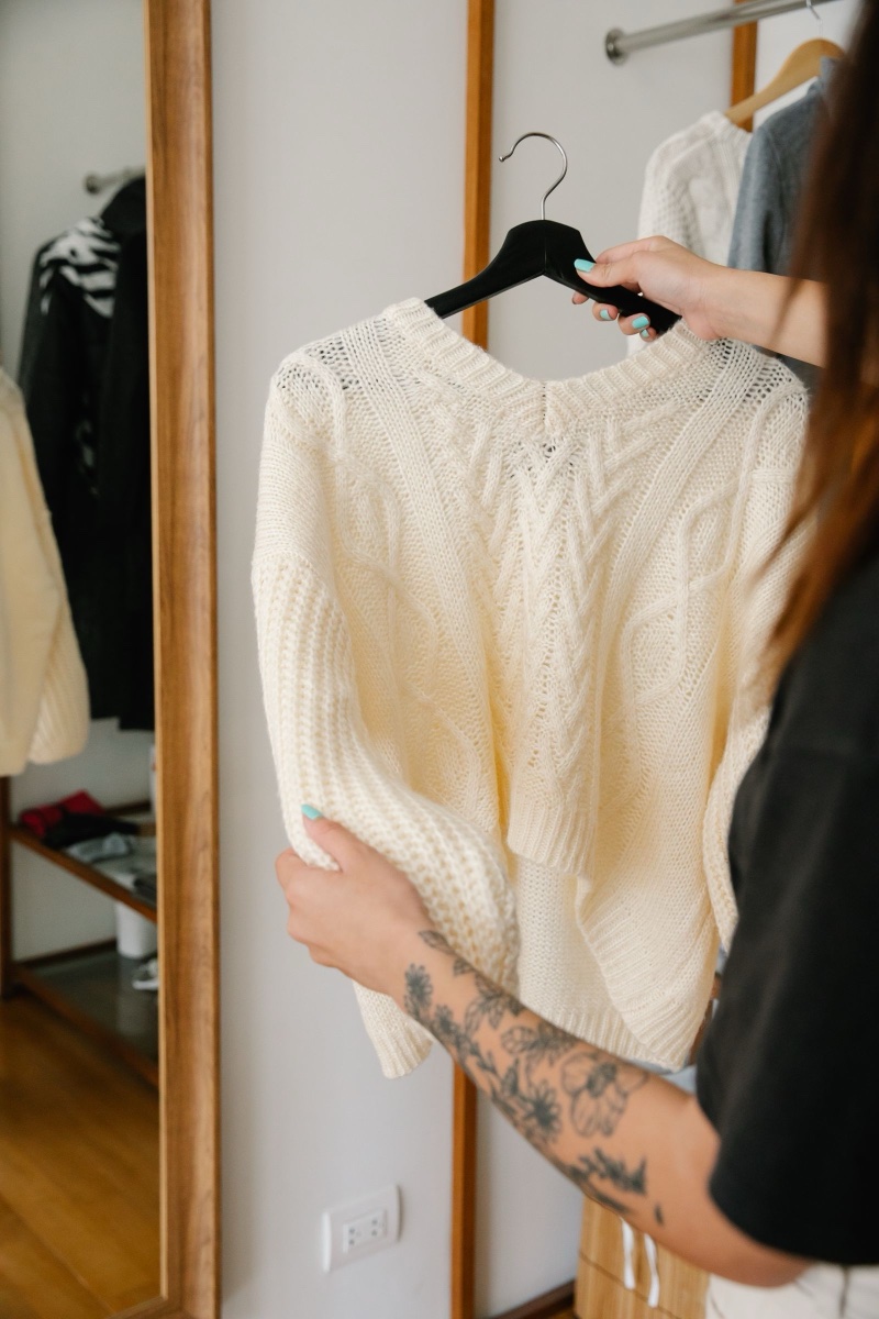 A person holding a cream cable-knit jumper on a black hanger in front of a full-length mirror beside a clothes rail