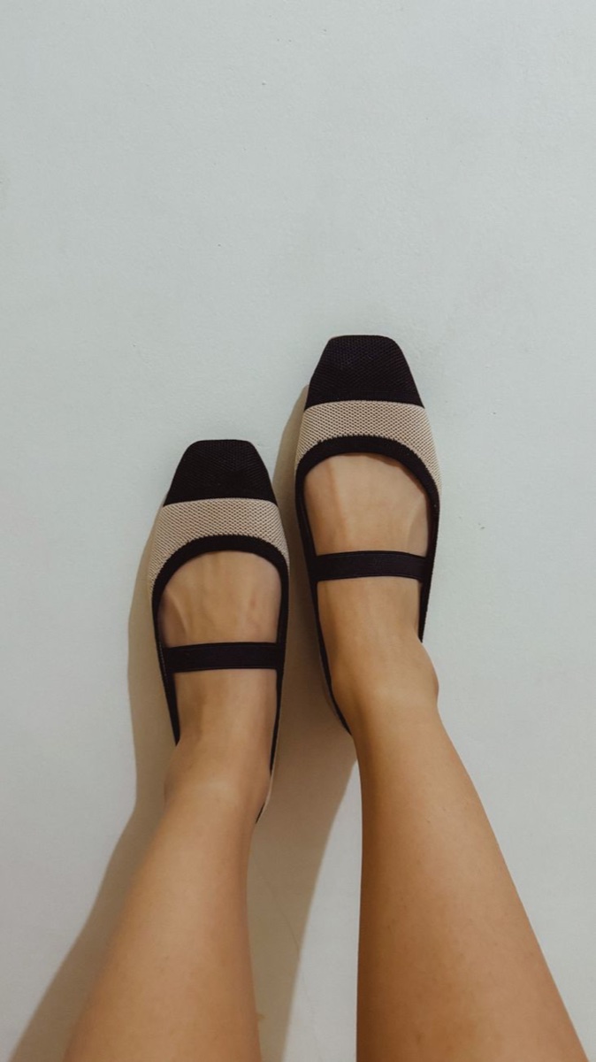 Two-tone knit Mary Jane flats in black and cream with elastic straps, worn bare-legged against a white wall