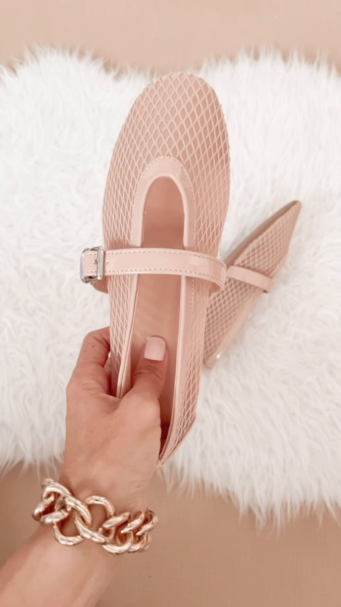 A pale pink fishnet Mary Jane ballet flat with a patent strap and silver buckle, held up in one hand