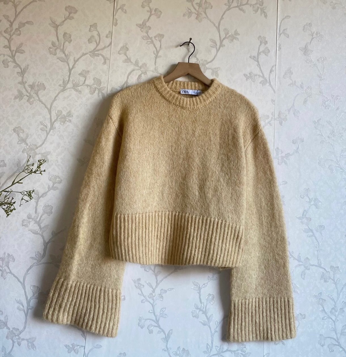 A pale yellow fuzzy Zara sweater on a wooden hanger against floral wallpaper, listed by a resale shop