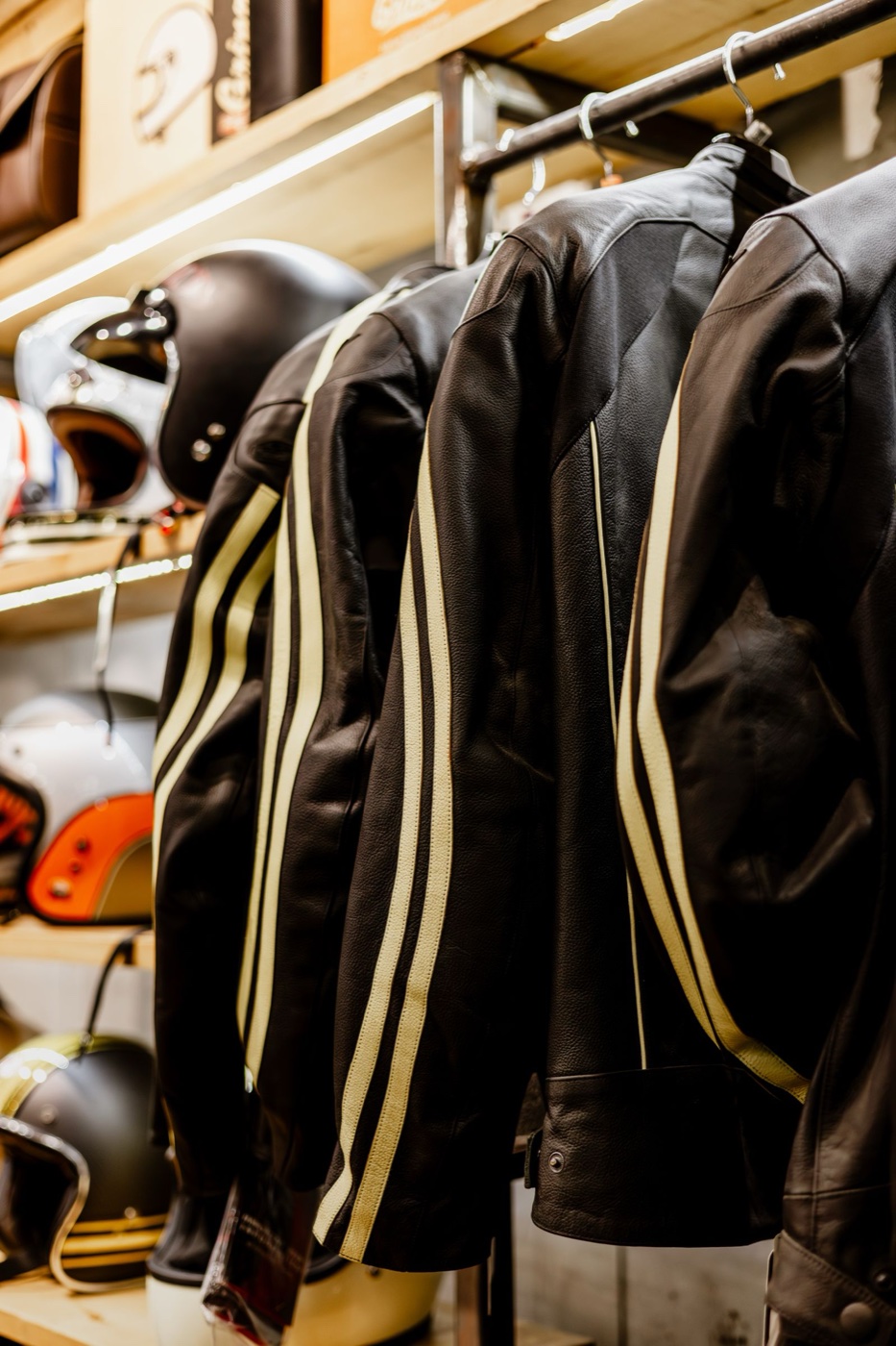 Black leather motorcycle jackets with cream racing stripes hanging in a motorcycle gear shop, helmets on shelves behind