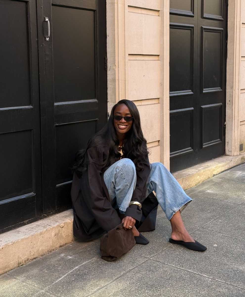 Woman in a dark trench and light wide-leg jeans crouching on a stone doorstep in black flat pointed mules, brown suede bag on the ground