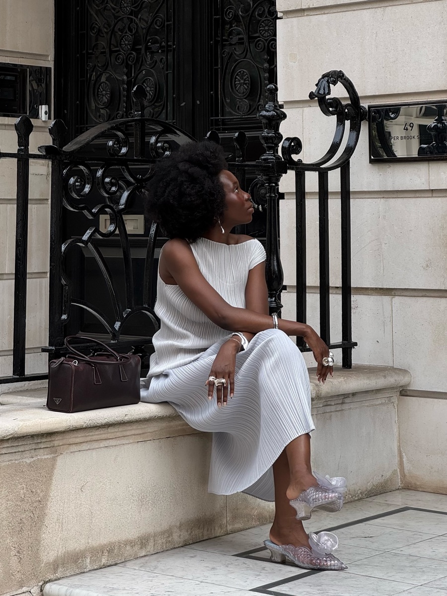 Woman in a pale grey pleated dress sitting on a stone ledge by wrought-iron railings, wearing clear sculpted kitten-heel mules