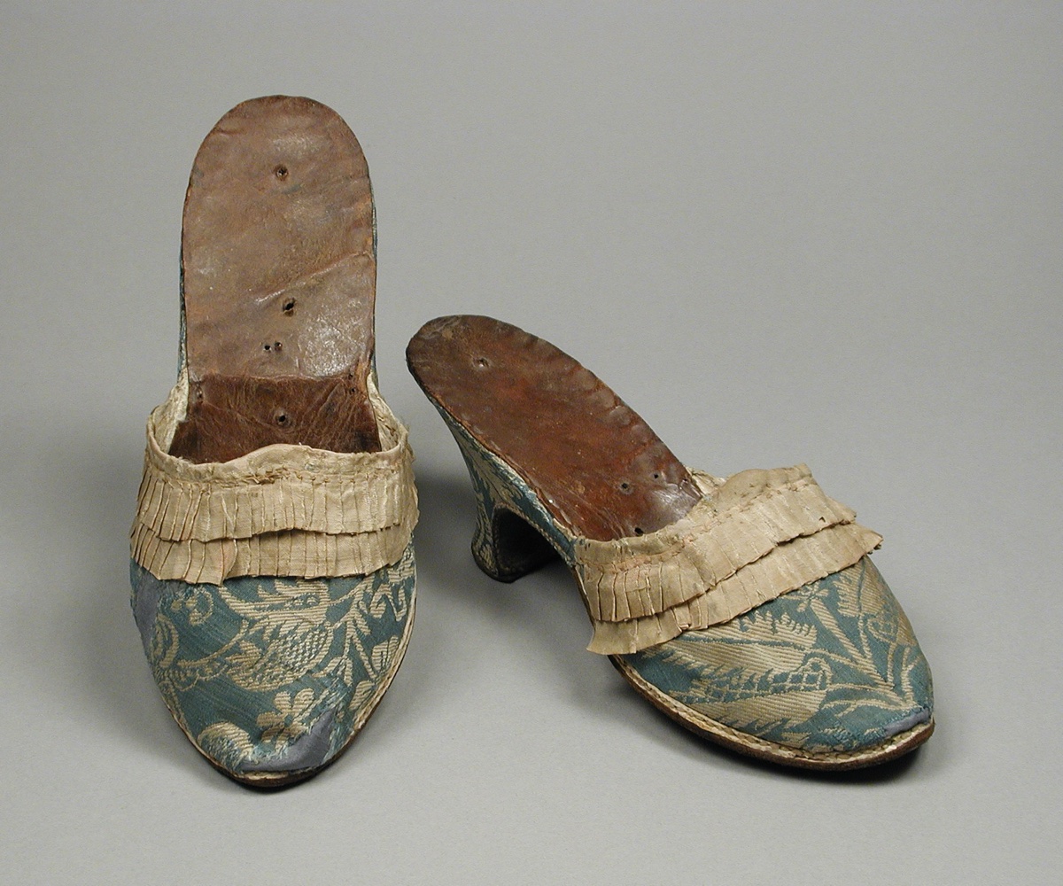 A pair of blue and cream silk brocade heeled mules from the 1770s with worn brown leather insoles, photographed against a gray museum background