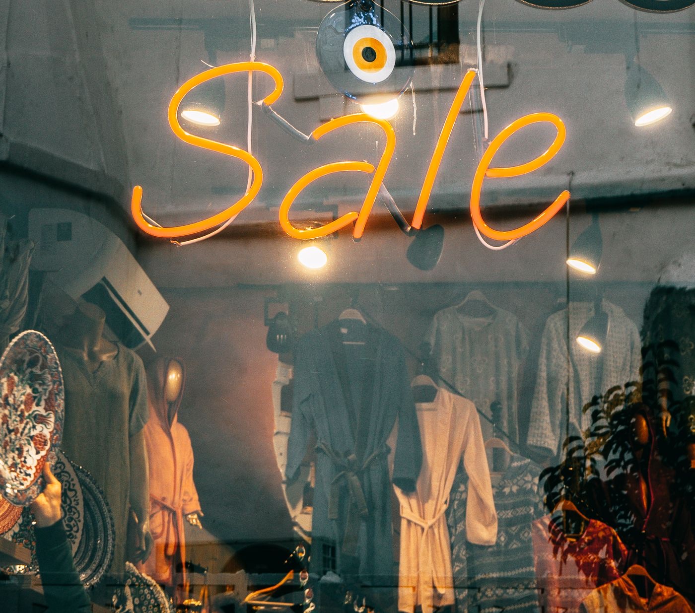 An orange neon script reading Sale glowing in a shop window at dusk, with robes and hanging garments visible behind the glass