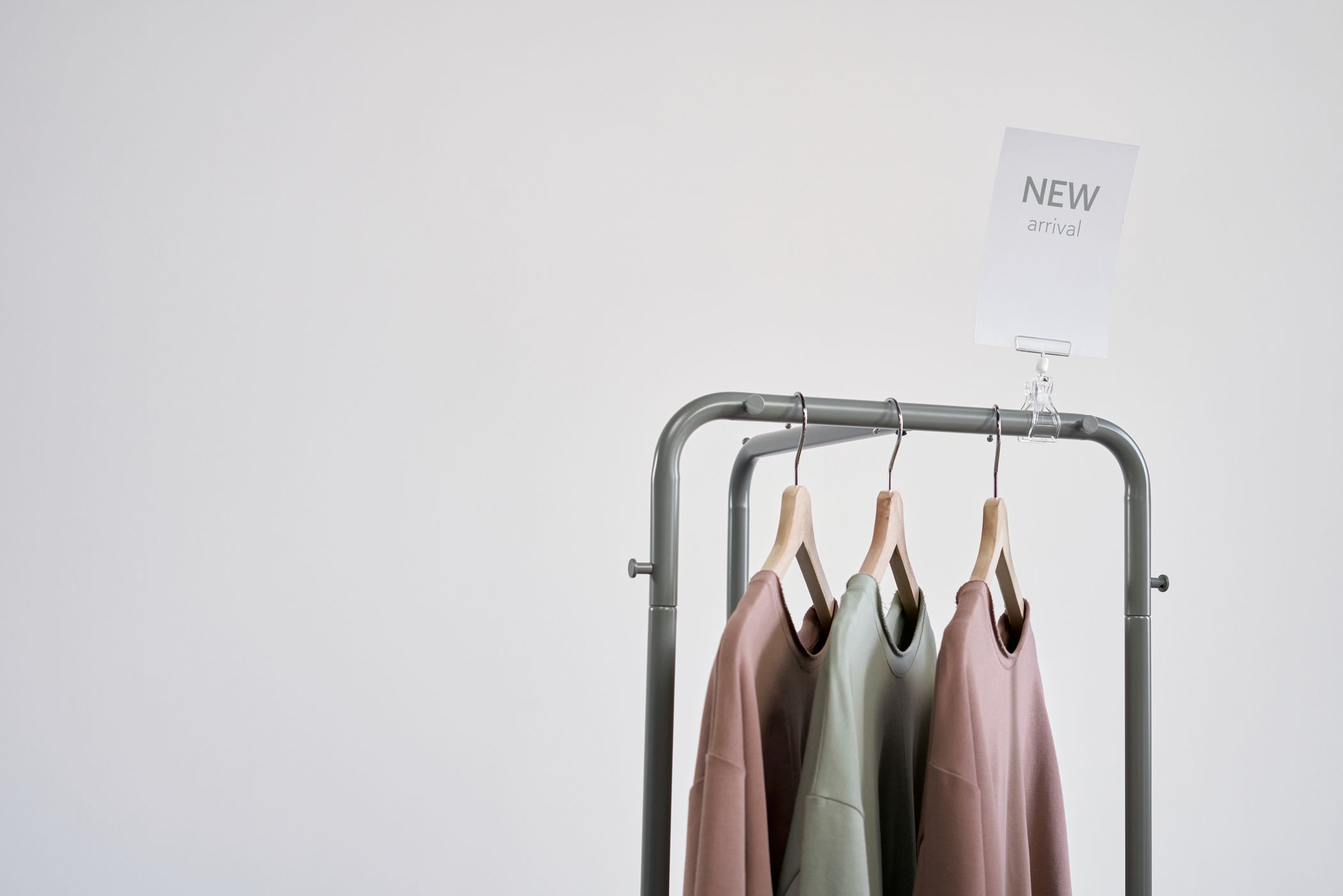 A grey clothing rail against a white wall holding three plain sweatshirts in pink, sage and dusty rose under a small card reading NEW arrival