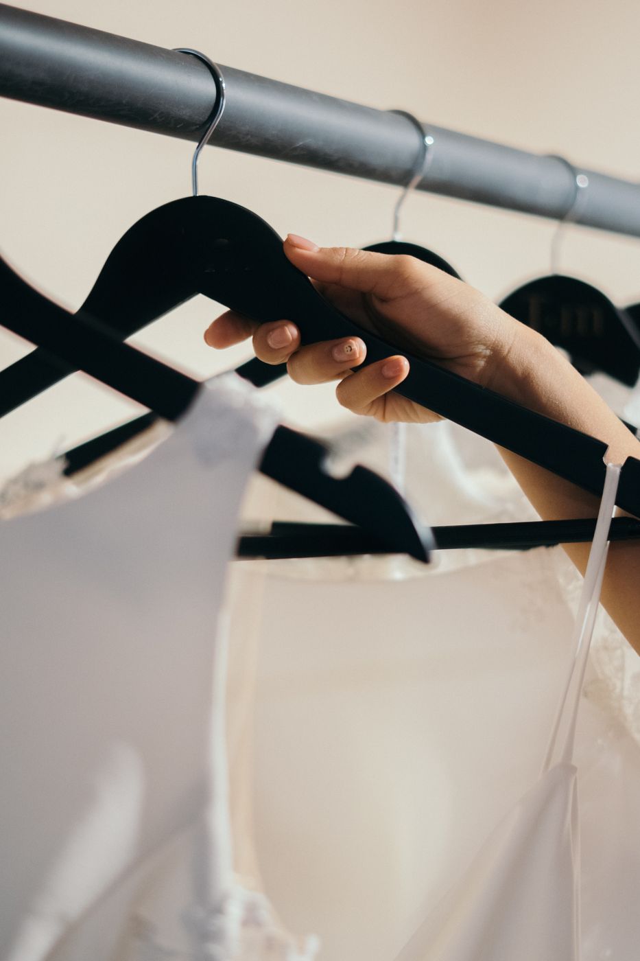 A hand reaching up to pick a black hanger holding a white dress from a rail of white garments in soft indoor light
