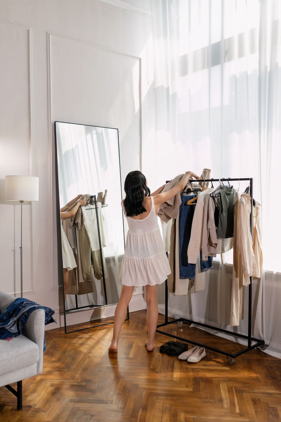 A woman seen from behind in a white dress arranging clothes on a freestanding rack beside a full-length mirror in a sunlit room with parquet flooring