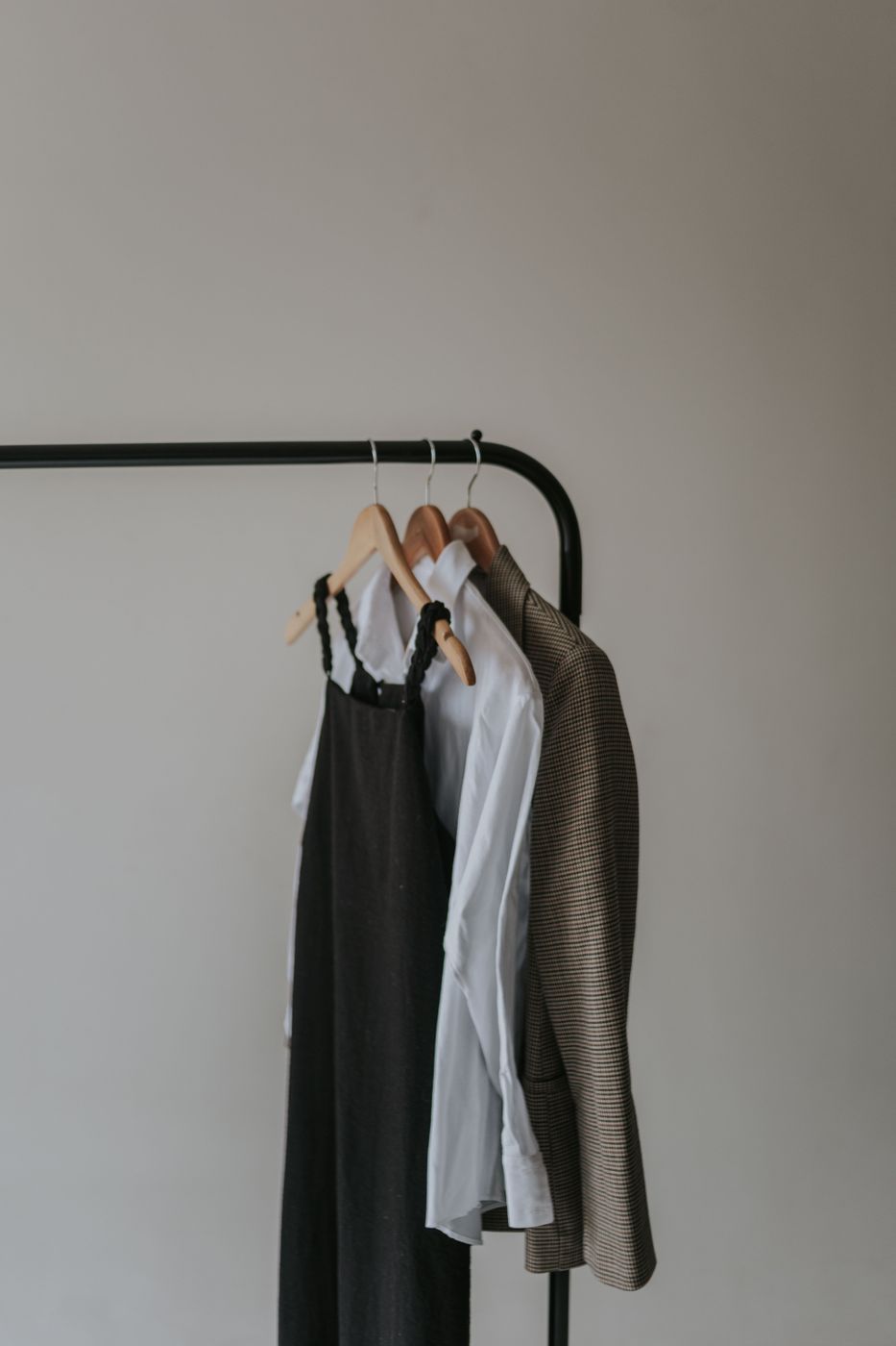 Three garments on wooden hangers, a dark slip dress, a white shirt and a houndstooth blazer, hanging from a black clothing rack against a bare wall