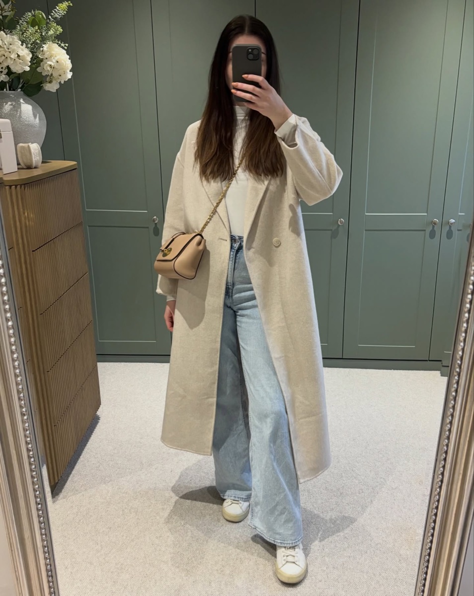 Mirror photo of a long oatmeal double-faced wool coat worn open over a white top and light-wash wide jeans, in a dressing room with sage green wardrobes