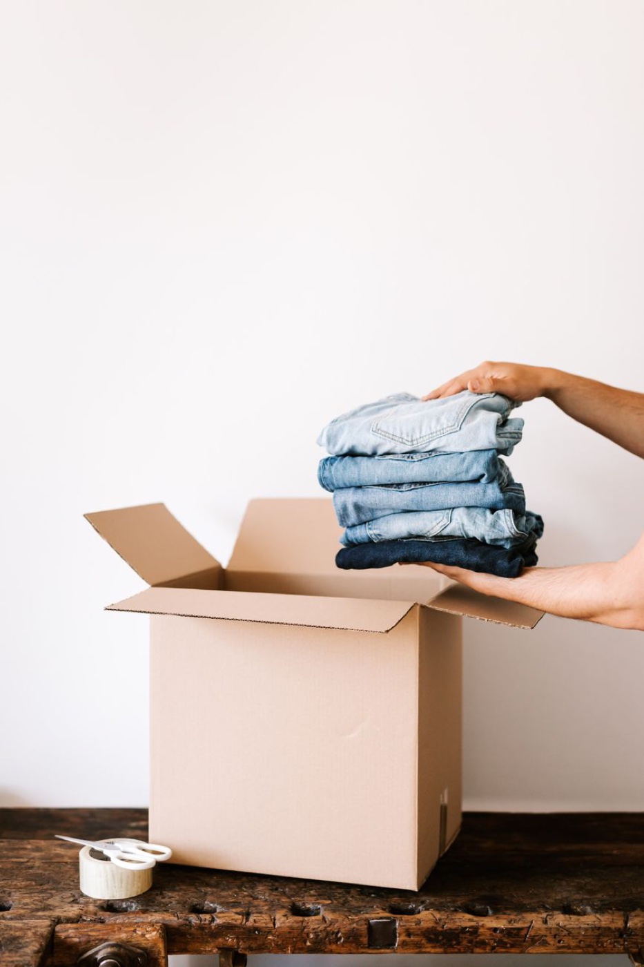 A pair of hands lowering a stack of five folded jeans into an open cardboard box on a wooden table