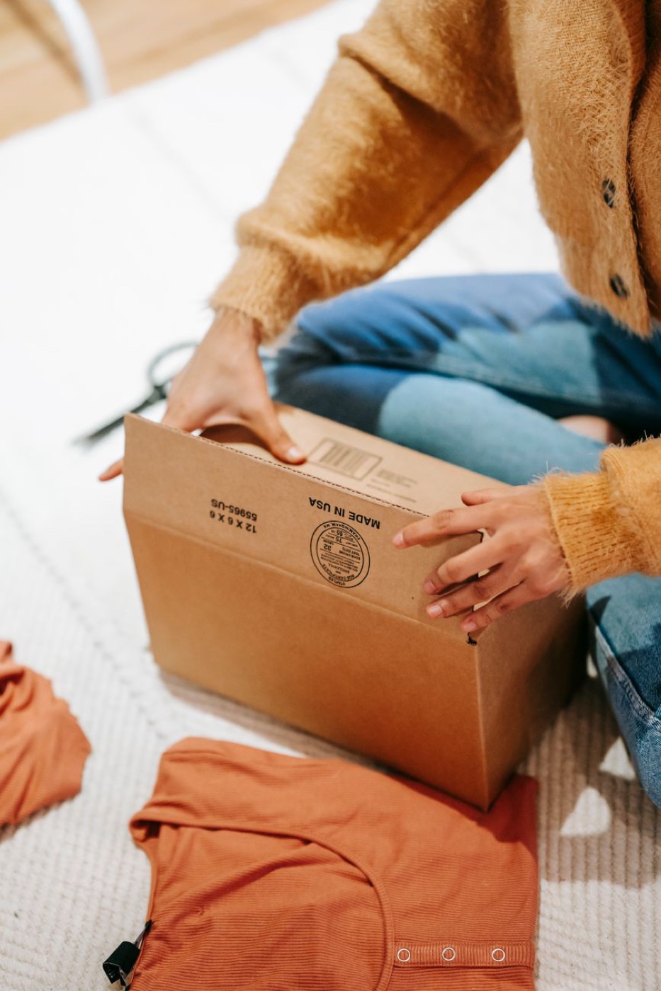 A person in a mustard cardigan closing a small cardboard box on a rug with folded orange knitwear beside it