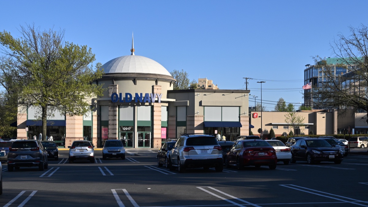 Old Navy store with a white domed roof and blue lettering behind a parking lot of cars at a suburban shopping center, early evening
