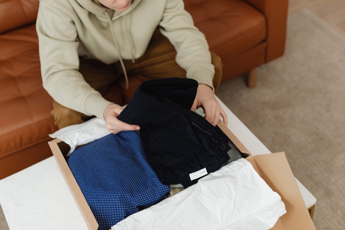 A person unpacking folded navy trousers and a polka-dot shirt from a cardboard delivery box on a coffee table