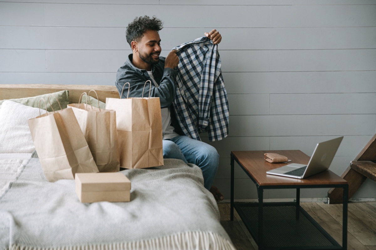 A man sitting on a bed holding up a plaid shirt beside paper shopping bags and an open laptop