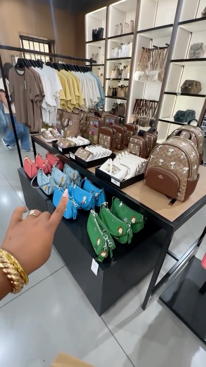 Display table inside a Coach Outlet stacked with small Snoopy-print bags in coral, blue and green, a shopper's hand pointing at one