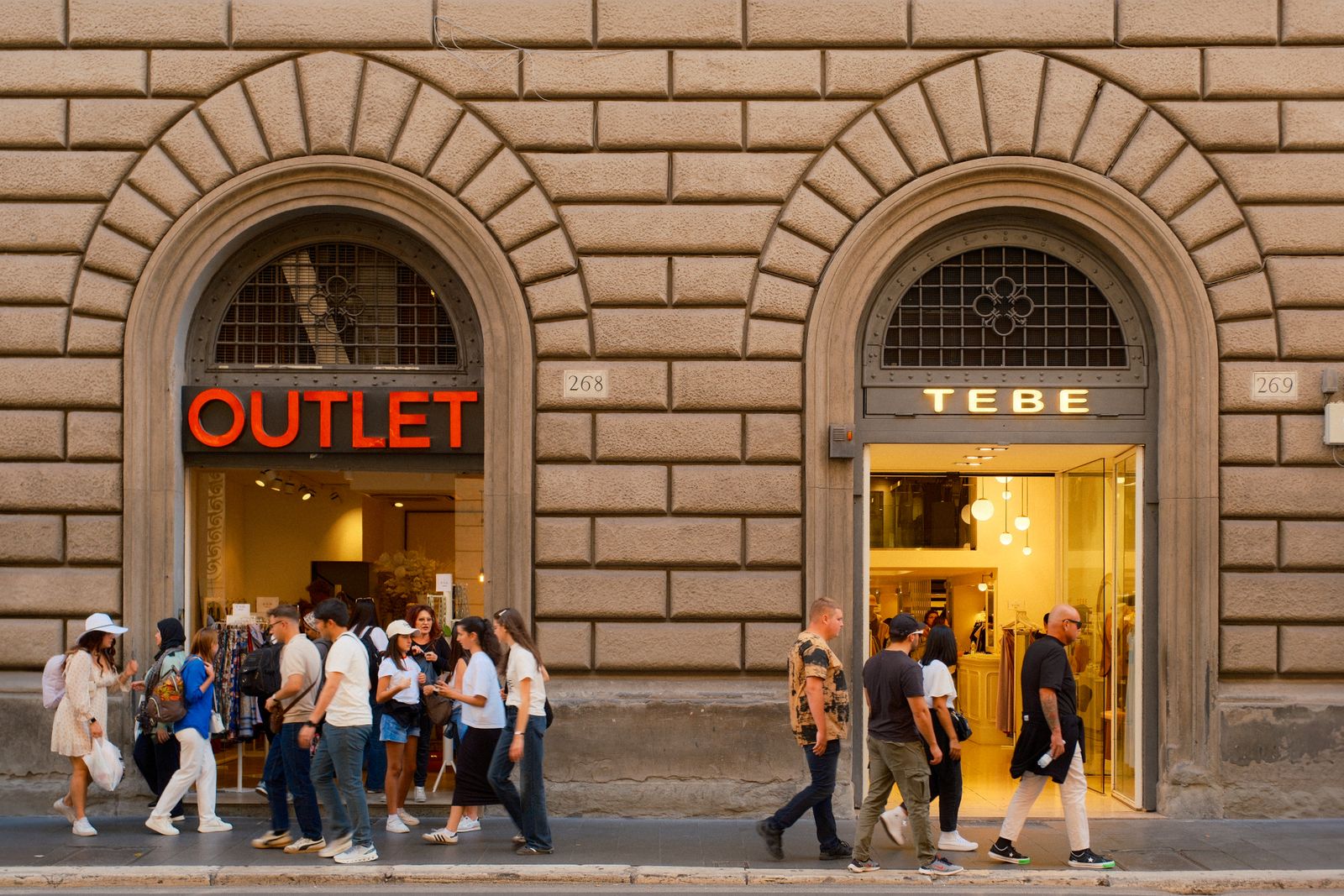 Shoppers walking past a stone storefront with a red OUTLET sign over an arched doorway in Florence