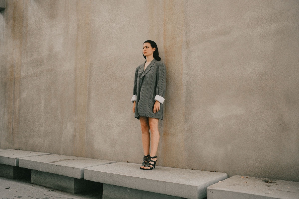 A woman stands on a concrete bench wearing an oversized gray Prince of Wales check blazer with the white cuffs rolled back