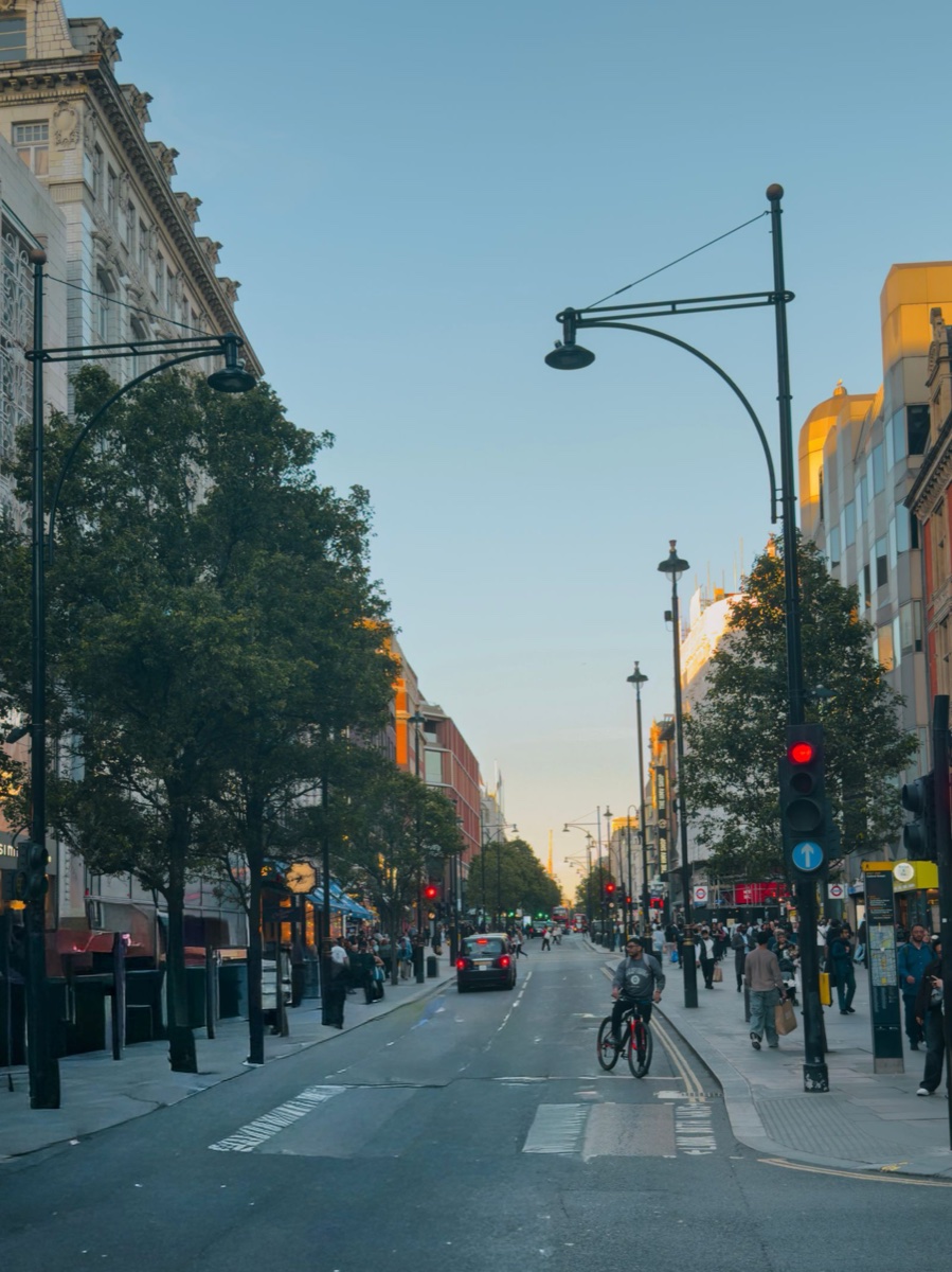 Oxford Street in London at dusk, shop lights coming on, pedestrians with shopping bags on the right pavement and a cyclist in the road