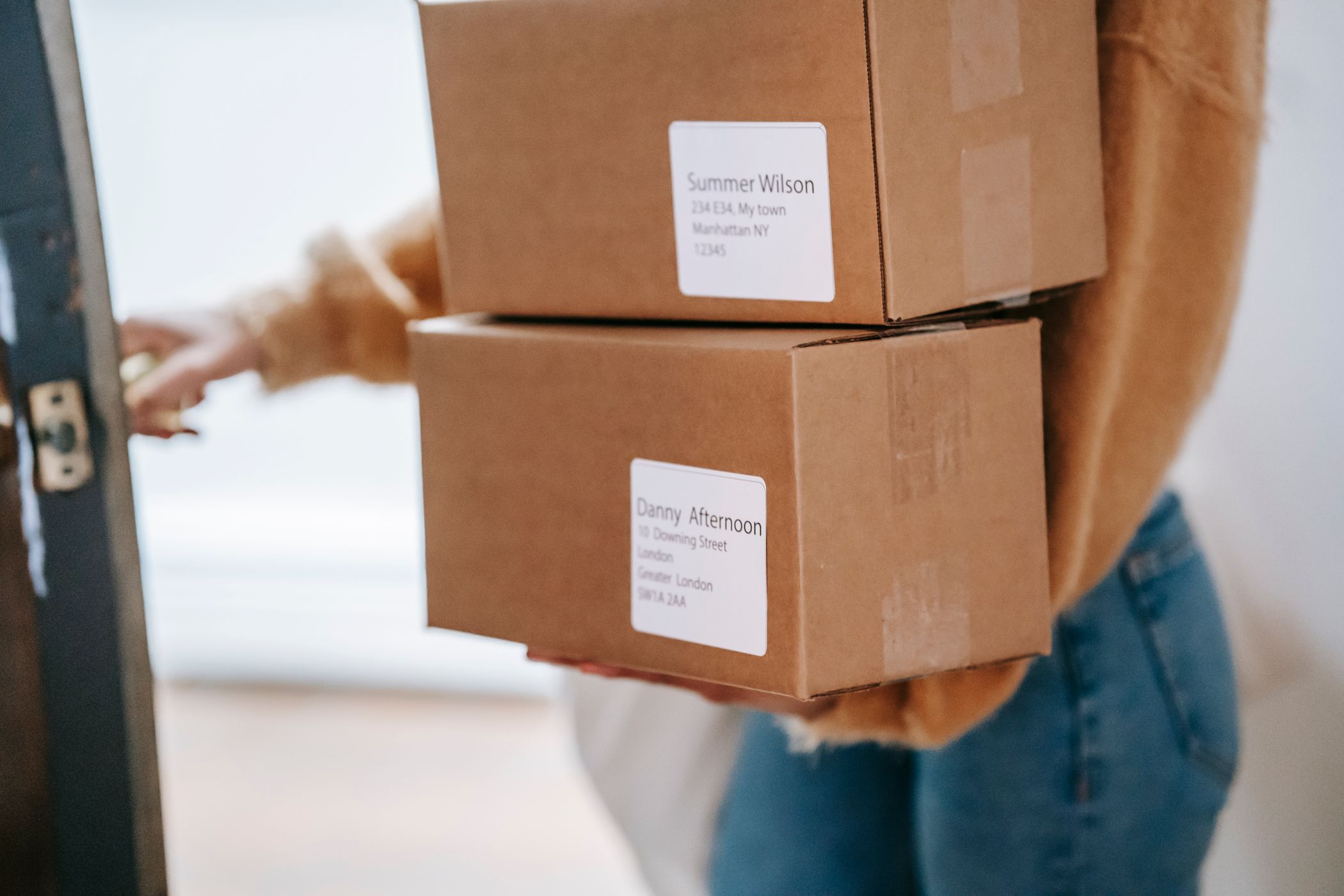 A person in a mustard sweater carrying two cardboard parcels with printed address labels through a doorway